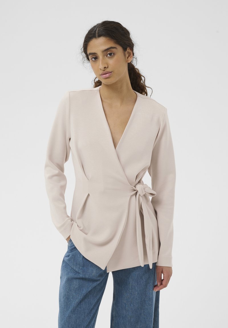 Jeune femme aux cheveux bruns bouclés portant un blazer cache-cœur beige pâle noué à la taille et un jean en denim bleu, debout devant un fond blanc.