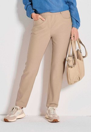 Beige Stretchhosen mit glatter Textur, Seitentaschen und kontrastierenden Nähten, kombiniert mit hellen Sneakers und einer passenden Handtasche.