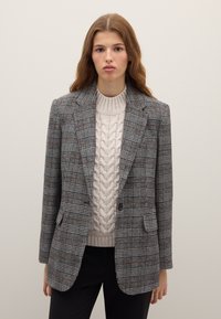 Grijze geruite blazer met gestructureerde schouders, twee voorzakken en een enkele knoopsluiting, gedragen over een kabelgebreide beige trui.