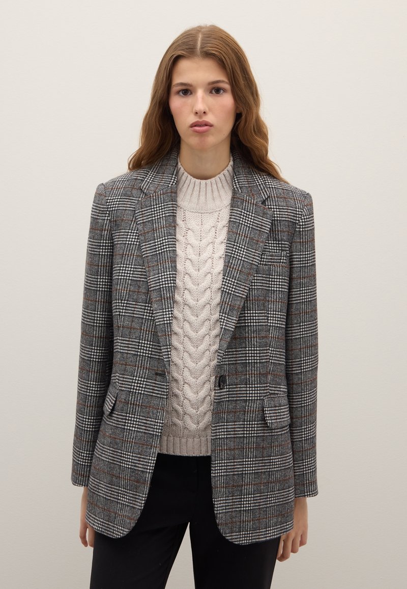 Grijze geruite blazer met gestructureerde schouders, twee voorzakken en een enkele knoopsluiting, gedragen over een kabelgebreide beige trui.