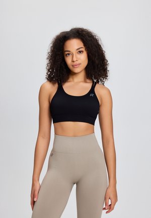 Ung kvinde med krøllet hår iført en sort sports-bh og beige leggings med høj talje, stående mod en ensartet lys baggrund.