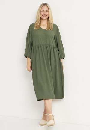 Grünes, knielanges Kleid aus strukturiertem Stoff mit V-Ausschnitt, gerafftem Taillenbereich und drei Viertel langen Ärmeln. Kombiniert mit beigen Espadrilles.