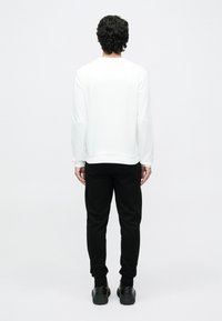 Göndör, sötét hajú férfi, fehér hosszú ujjú inget, fekete nadrágot és fekete cipőt viselve, háttal áll egy egyszerű háttér előtt.