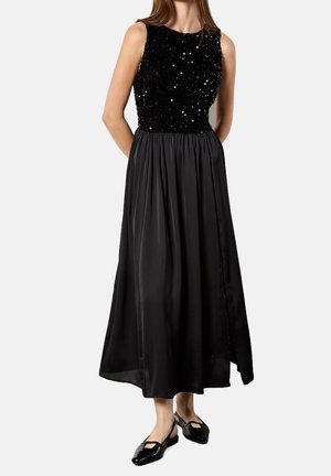 Femme portant une robe noire sans manches avec un corsage orné de sequins et une jupe fluide en satin, assortie à des chaussures plates noires.
