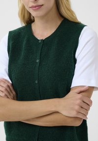 Grüne Strickweste mit rundem Ausschnitt, die sechs Knöpfe auf der Vorderseite hat und über einem weißen kurzärmligen Hemd getragen wird.