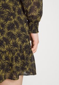 Gros plan d'une robe vert olive avec un motif floral abstrait noir, montrant une manche à manchette élastique et un ourlet froncé à côté d'une main.