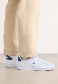 Sneaker bianco realizzato in materiale liscio, con un accento verde sul tallone, dotato di dettagli minimi e un semplice design con lacci.