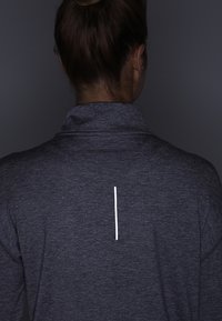 Maglione grigio scuro a maniche lunghe con colletto alto. Presenta una striscia verticale bianca sul retro. Realizzato in un tessuto strutturato e elasticizzato.