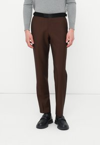 Bărbat îmbrăcat în pantaloni maro personalizați cu detalii negre, curea neagră, pantofi negri și un top aderent gri cu mânecă lungă, stând pe un podea albă.