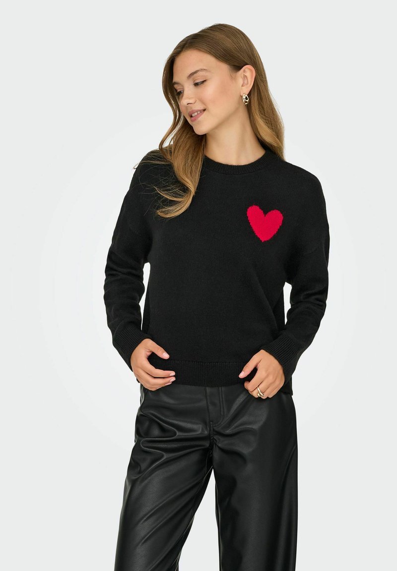 Maglione nero lavorato a maglia con un cuore rosso sul petto. Presenta un collo rotondo e polsini e orlo a costine. Abbinato a pantaloni di pelle nera.