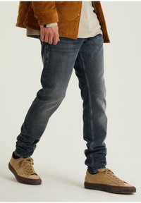 Avslappnad outfit med mörkblå jeans i slim fit, tillsammans med ljusbruna mockasneakers och en texturerad senapsgul kordjacka.