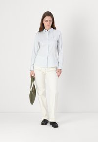 Camicia button-up a righe blu chiaro con maniche lunghe, abbinata a pantaloni avorio e una borsa verde. Scarpe nere completano il look.