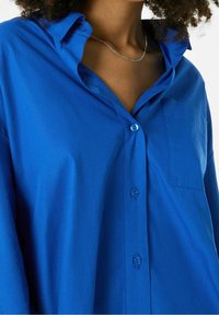 Camisa azul de botones con cuello, con un bolsillo. Hecha de tela suave con un corte holgado y botones brillantes.