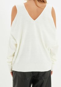 Pull blanc à manches longues avec un dos en V et des découpes ouvertes aux épaules. Présente un ourlet et des poignets côtelés, fabriqué en matériau doux et léger.