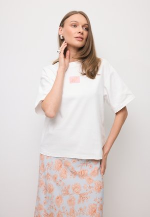 Kobieta w białej koszulce z krótkim rękawem z różowym logo i jasnoniebieskiej spódnicy w pomarańczowy wzór kwiatowy, pozująca z jedną ręką na szyi.