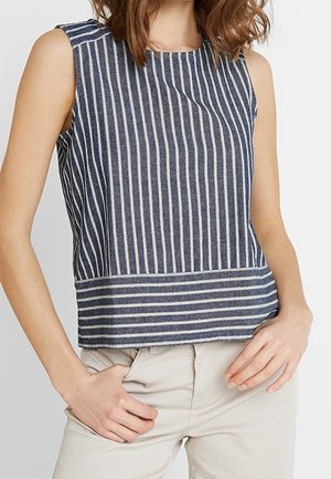 Vrouw draagt mouwloze donkerblauwe top met verticale witte strepen en beige broek, poseert met één hand in de zak.
