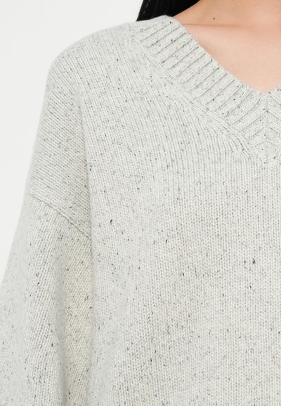 Vaalea harmaa neulepusero, jossa on V-pääntie, ribattu kaulus ja pilkullinen tekstuuri. Pusero tarjoaa rennon siluetin ja pehmeää materiaalia.