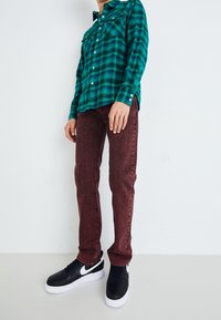 Camisa de franela a cuadros verde, vaqueros rojos oscuros con un acabado texturizado y zapatillas Nike negras con suelas blancas. Conjunto casual con detalles de botones.