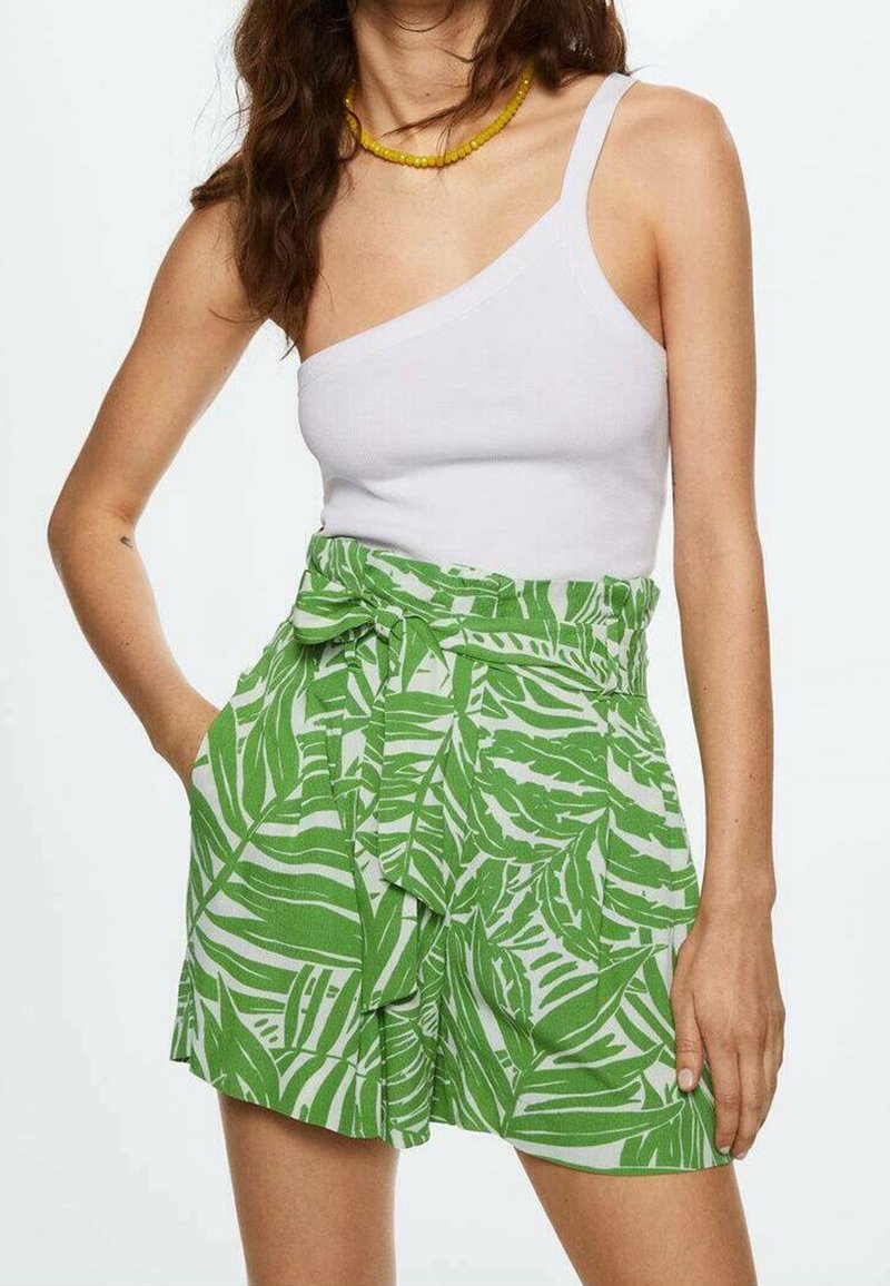 Femme portant un top blanc asymétrique et un short vert taille haute avec un imprimé feuilles tropicales blanches, mains dans les poches, collier de perles jaunes.