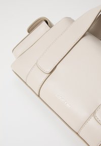Bež torba Calvin Klein od glatke kože, širokih traka i minimalističkog dizajna. Sadrži suptilan logo sa strane.