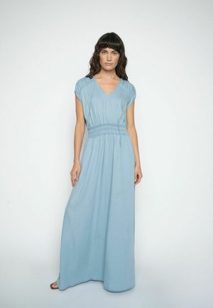 Kvinde med mørkt hår iført en lyseblå, ærmeløs maxikjole med samlet ærmekant og indsnøret talje, stående foran en ensfarvet baggrund.