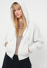 Felpa grigia con zip, dotata di un ampio cappuccio, tasche frontali e cordini bianchi a contrasto; morbido al tatto e vestibilità rilassata.