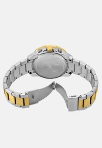 Sidabrinė ir aukso atspalvių riešo laikrodis su nerūdijančio plieno juosta, turintis apvalų korpusą, tekstūruotą rėmelį ir saugų užsegimą.