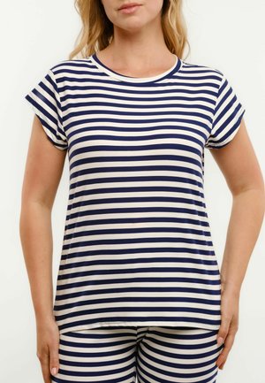 Nő tengerészkék és fehér vízszintes csíkos rövid ujjú felsőt és hozzá illő nadrágot visel, egyszínű világos háttér előtt áll.