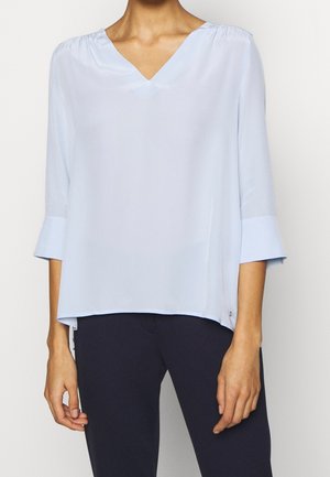 Kvinde iført lyseblå V-halsbluse med trekvartlange ærmer og marineblå bukser, stående med hænderne afslappet langs siderne.