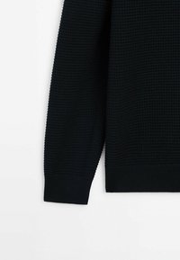 Pull en maille noir à manches longues avec poignets et ourlet côtelés, présentant un motif texturé sur fond blanc.