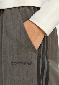 Ruka u džepu sivih Adidas hlača s crnim prugama i logom, nosi se s bijelom majicom dugih rukava.