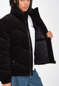 Schwarz corduroy Pufferjacke mit hohem Kragen, Reißverschluss und elastischen Bündchen. Das Innere verfügt über ein elegantes Futter und eine Tasche. Begleitet von einem weißen T-Shirt.