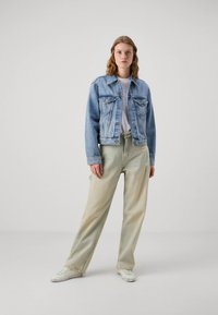 Gaiši zilas krāsas džinsa jaka, zem tās balta T-krekls, kā arī izbalējušas zaļas platās bikses ar izšuvuma detaļām, kas kopā ar baltām teniskām veido komplektu.