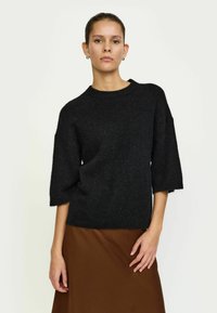 Melna adīta džemperis ar noapaļotu kakla izgriezumu un īsām, platām piedurknēm. Tekstūra izskatās maiga un nedaudz pūkaina, kopā ar brūnu svārku.