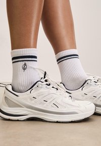 Baskets blanches avec des détails en maille, accents noirs et une bande argentée, associées à des chaussettes blanches côtelées ornées de rayures marines.