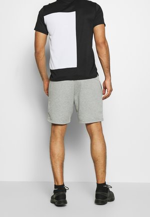 Mann trägt schwarz-weißes kurzärmeliges Hemd, graue Shorts, schwarze Socken und schwarze Turnschuhe, steht mit dem Gesicht zur weißen Wand.