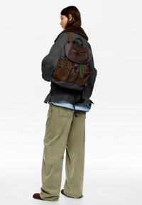 Rjav in siv nahrbtnik z več zadrgami, vzorcem karo, nastavljivimi trakovi in zgornjim pokrovom. Nosijo ga oseba v ohlapnih oblačilih.