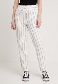 Person som bär vita byxor med tunna vertikala grå ränder, dragkedja i midjan, svart topp och svart-vita sneakers, stående på ljus bakgrund.