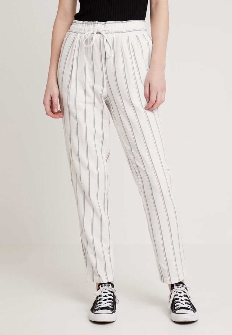 Person som bär vita byxor med tunna vertikala grå ränder, dragkedja i midjan, svart topp och svart-vita sneakers, stående på ljus bakgrund.