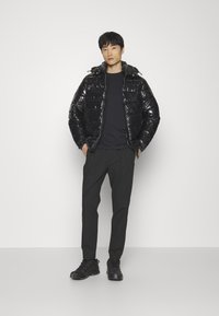Zwarte gewatteerde jas met een glanzende afwerking, voorzien van een capuchon en een voorrits, gecombineerd met zwarte broek en zwarte sportschoenen.
