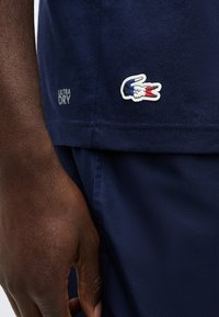 Χέρι με σκούρο δέρμα δίπλα στο τελείωμα μπλε σκούρου πουκάμισου με πολύχρωμο λογότυπο κροκόδειλου και κείμενο "Ultra Dry", σετ με αντίστοιχες μπλε σκούρες σορτς.