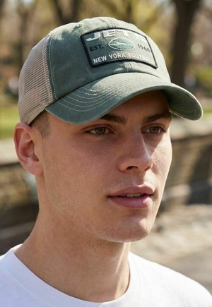 Junger Mann trägt grüne und beige Baseballkappe aus Mesh mit dem Aufnäher "Jets New York Football" im Freien bei weichem Sonnenlicht.