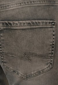 Tmavě šedá džínová kapsa s přehnutým poklopem, zdobená prošitým okrajem a stříbrným knoflíkem. Textura je mírně opotřebovaná.