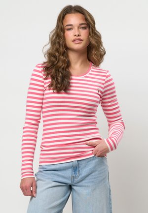 Mladá žena s vlnitými vlasmi, oblečená v ružovo-bielom pruhovanom tričku s dlhým rukávom a svetlomodrých džínsoch, stojaca s rukou v vrecku.