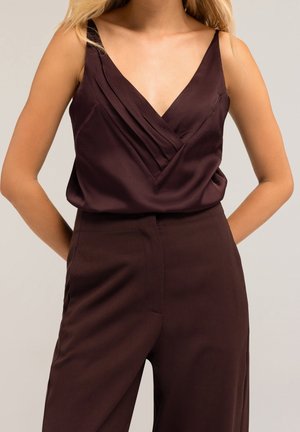 Vrouw draagt een mouwloze donkerpaarse satijnen top met V-hals, gecombineerd met bijpassende hoge taille broek, staat met handen achter haar rug.