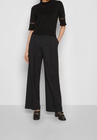 Top negro ajustado con bordes festoneados y mangas cortas; combinado con pantalones negros de pierna ancha de cintura alta y mocasines negros de tacón grueso.