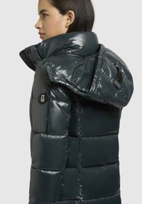 Pufota jaka tumši zaļā krāsā, spīdīgā materiālā, ar kapuci, ar rāvējslēdzēja kabatām un mazu zīmola logo uz krūšdaļas.