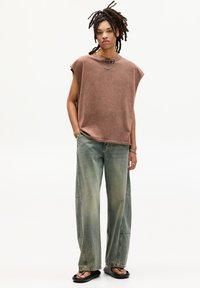 Un top oversized marron sans manches, un jean large bleu clair, et des tongs noires. Collier en chaîne avec un pendentif notable. Texture de tissu lisse.