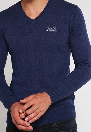 Mand iført en tætsiddende marineblå V-hals sweater med hvidt "SuperDry"-logo på brystet, hænder samlet foran, sorte bukser synlige.