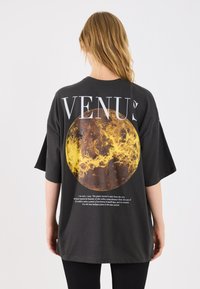 T-shirt oversize nera con una grande illustrazione gialla del pianeta Venere e testo descrittivo stampato sul retro.